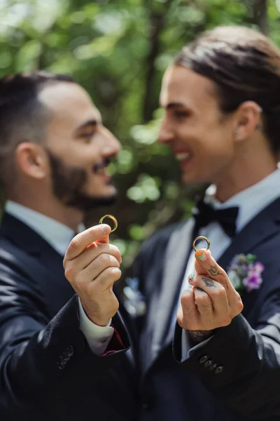 Happy and blurred gay men holding wedding rings in hands and looking at each other — Stock Photo