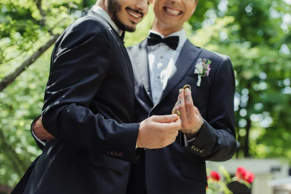 Partial view of happy gay couple in formal wear holding golden wedding rings in hands — Fotografia de Stock