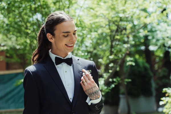 Happy tattooed man with golden wedding ring on finger smiling while adjusting floral boutonniere — Stock Photo