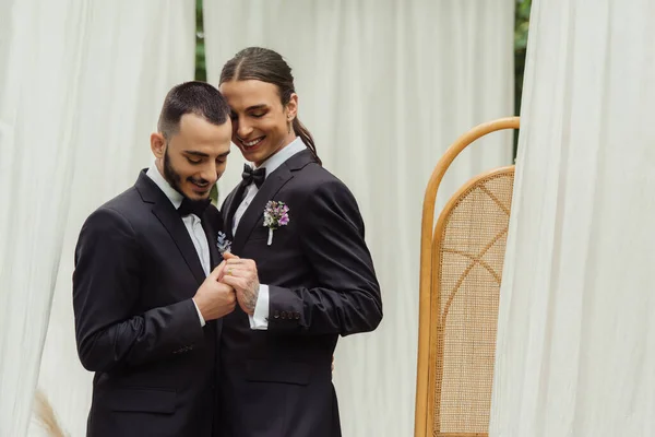 Feliz pareja gay en trajes con boutonnieres tomados de la mano en el día de la boda - foto de stock