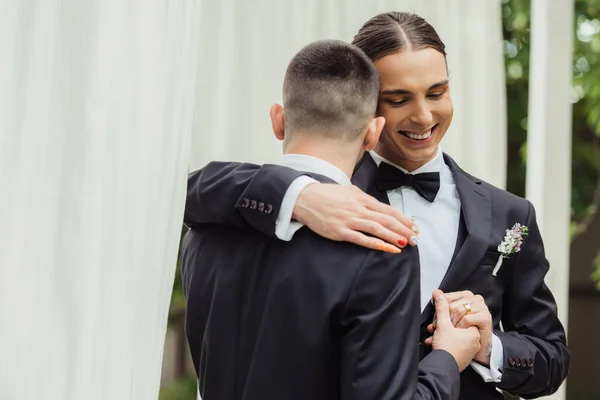 Glückliches homosexuelles Paar in Anzügen, Händchen haltend und sich umarmend am Hochzeitstag — Stockfoto