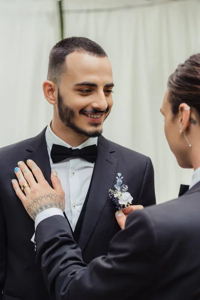 Tattooed gay man adjusting boutonniere on suit of happy bearded groom - foto de stock