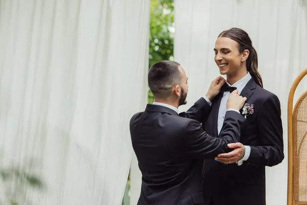 Gay man adjusting bow tie on suit of happy groom in formal wear — Stock Photo