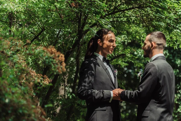 Happy gay newlyweds in formal wear with boutonnieres holding hands in green park — Stock Photo