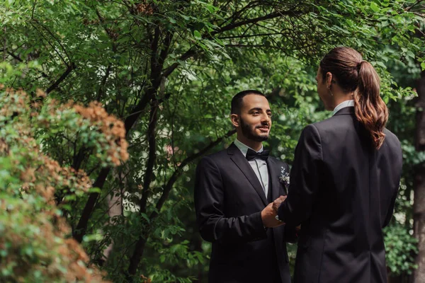 Счастливые молодожёны-геи в формальной одежде смотрят друг на друга и держатся за руки в парке — стоковое фото