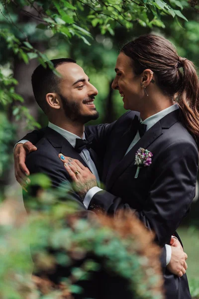 Seitenansicht von fröhlichen homosexuellen Brautpaaren in formeller Kleidung, die sich im grünen Park umarmen — Stockfoto