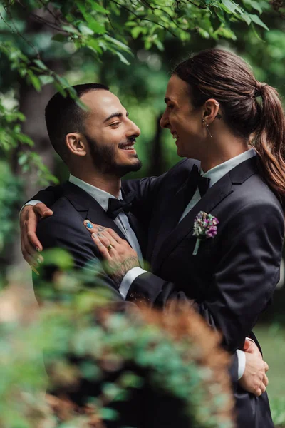 Side view of joyful gay newlyweds in formal wear hugging in green park — Stockfoto