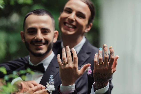 Blurred and happy gay newlyweds in formal wear with boutonnieres showing golden rings on fingers — Stock Photo