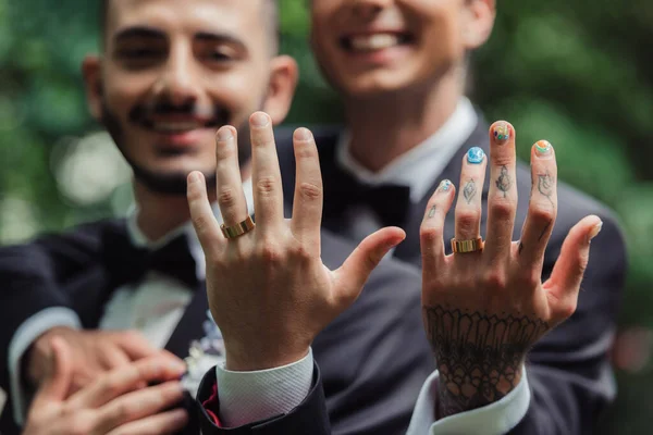 Blurred and cheerful gay newlyweds in formal wear with boutonnieres showing golden rings on fingers — Photo de stock