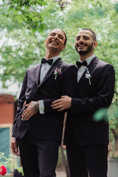 Low angle view of happy gay newlyweds in formal wear with boutonnieres and golden rings — Fotografia de Stock