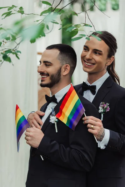 Happy gay newlyweds in formal wear with boutonnieres holding lgbt flags — Stock Photo