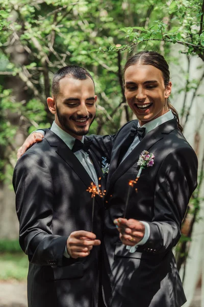 Cheerful gay newlyweds in formal wear with boutonnieres holding sparklers in green park - foto de stock