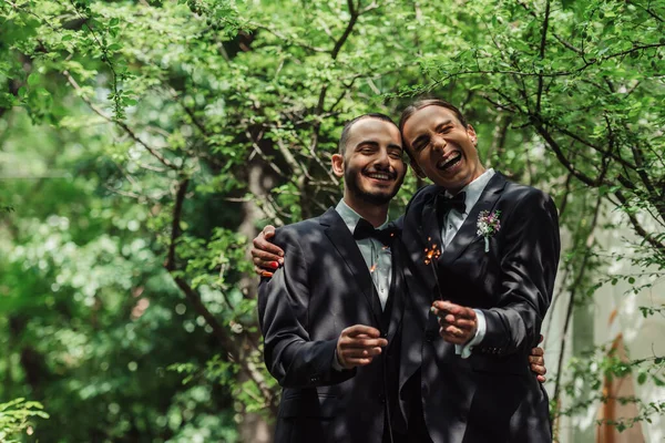 Happy gay newlyweds in formal wear holding sparklers in green park — Stockfoto