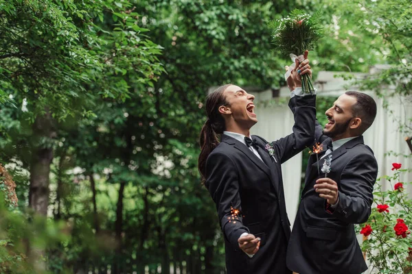 Excited gay man in formal wear holding wedding bouquet and sparkler near groom in green park — Stockfoto