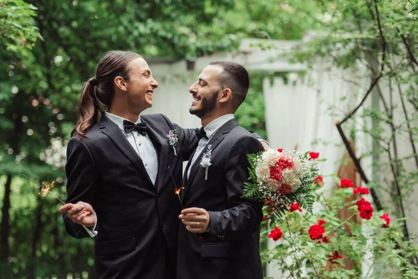 Pleased gay man in formal wear holding wedding bouquet and sparkler near groom in green park — Photo de stock