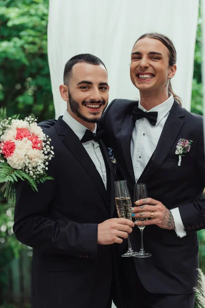 Feliz gay casal em ternos segurando copos de champanhe no casamento dia — Fotografia de Stock