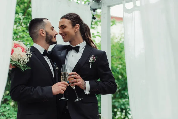 Alegre gay casal no ternos brindar copos de champanhe no casamento dia — Fotografia de Stock