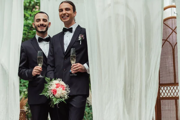 Alegre gay recém-casados no ternos segurando copos de champanhe no dia do casamento — Fotografia de Stock