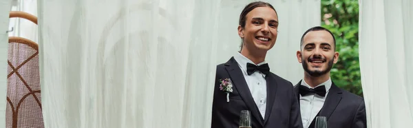 Happy gay couple in suits holding glasses of champagne on wedding day, banner — Stockfoto