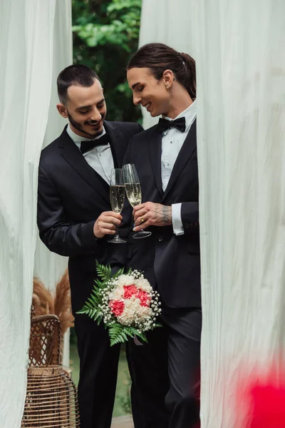 Smiling gay couple in suits clinking glasses of champagne on wedding day — Stock Photo