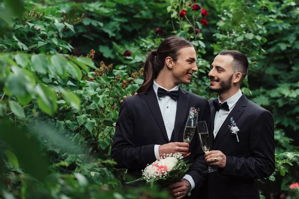 Feliz gay casal no ternos segurando copos de champanhe no verde parque — Fotografia de Stock