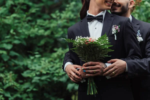 Corte vista de alegre gay casal em ternos segurando casamento buquê no verde parque — Fotografia de Stock