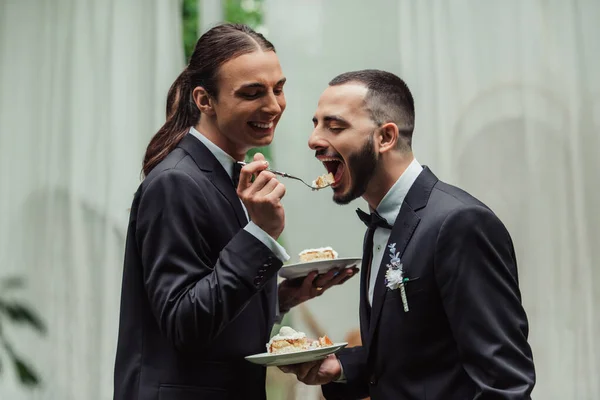 Cheerful gay man in formal wear feeding husband with wedding cake — Stockfoto