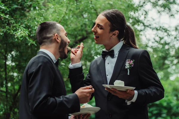 Tattooed gay man in formal wear feeding husband with wedding cake — Stock Photo