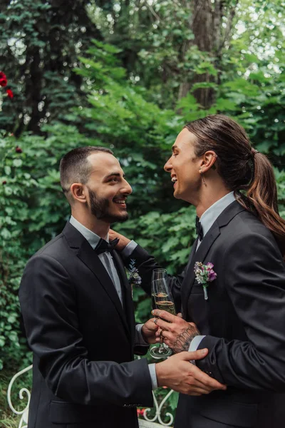 Vista laterale di felici sposi gay in abiti che tengono bicchieri con champagne il giorno del matrimonio — Foto stock