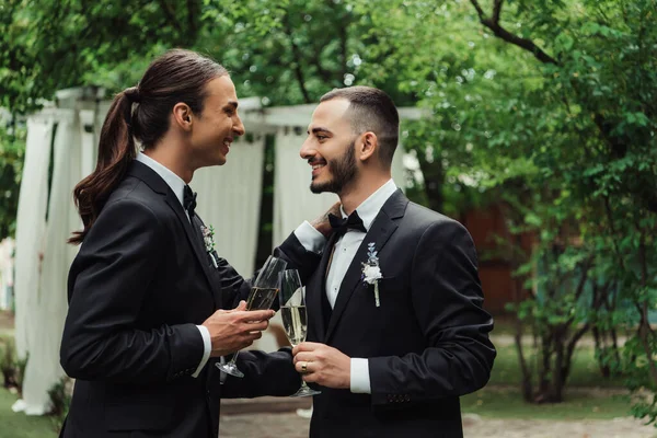 Vista laterale di allegri sposi gay in abiti che tengono bicchieri con champagne il giorno del matrimonio — Foto stock