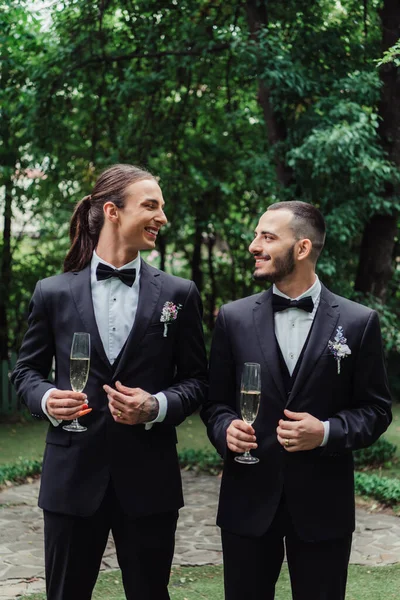 Pleased gay newlyweds in suits holding glasses with champagne on wedding day — Stock Photo