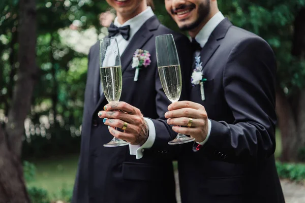 Corte vista de alegre gay recém-casados em ternos segurando copos com champanhe no dia do casamento — Fotografia de Stock