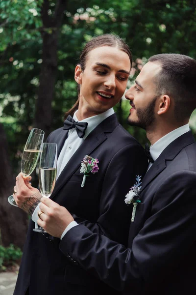 Happy gay newlyweds in suits holding glasses with champagne and looking at each other — Foto stock