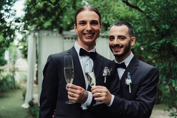 Веселые молодожёны-геи в костюмах, держащие бокалы с шампанским в день свадьбы — стоковое фото