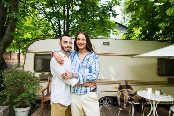 Happy gay couple smiling while hugging near travel van in forest — Foto stock