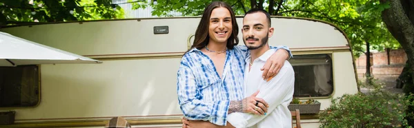 Щаслива гей-пара посміхається, обіймаючись біля фургона в лісі, банер — стокове фото
