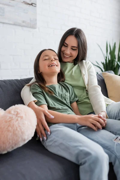 Happy brunette babysitter hugging cheerful kid while sitting on couch near soft pillow — Photo de stock