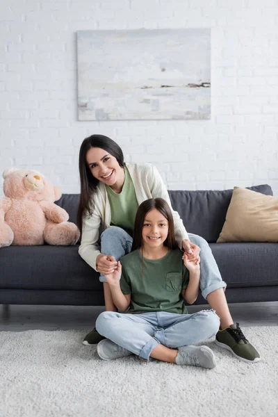 Glückliche Brünette Babysitter sitzt auf dem Sofa und hält Hände mit fröhlichen Mädchen — Stockfoto
