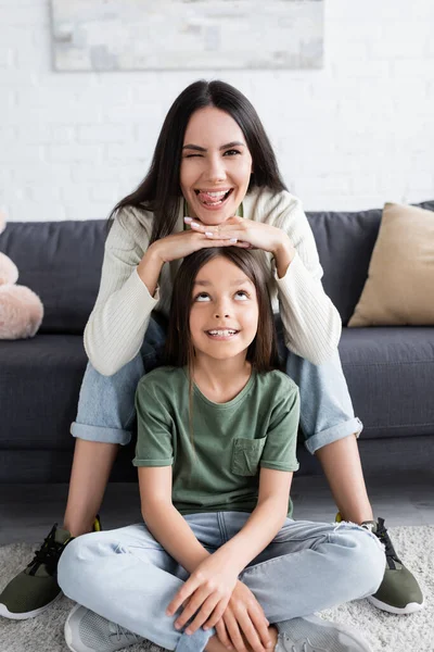 Heureux brunette baby-sitter collant sur la langue et assis sur canapé près enfant gai — Photo de stock