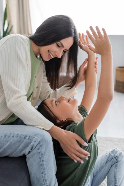 Happy brunette babysitter sitting on couch and looking at cheerful kid with outstretched hands — Stock Photo
