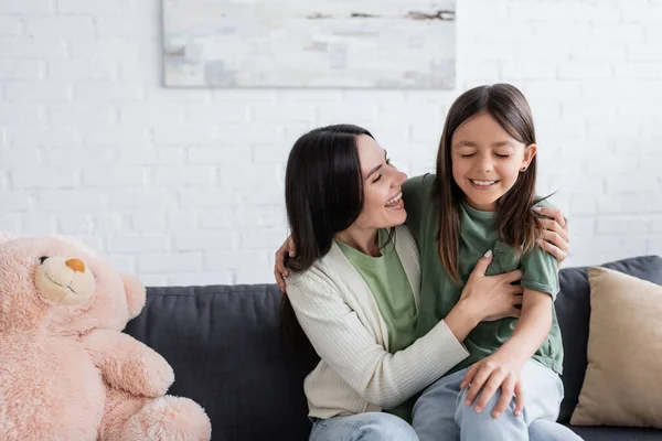 Aufgeregte Brünette Babysitter umarmt fröhliches Mädchen, während es auf der Couch sitzt — Stockfoto