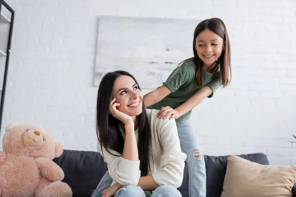 Playful girl standing behind happy babysitter resting on couch in living room — Fotografia de Stock