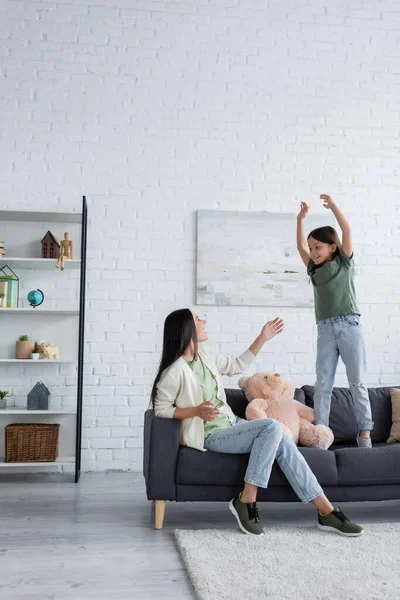 Amazed babysitter looking at excited girl gesturing while standing on sofa in living room - foto de stock