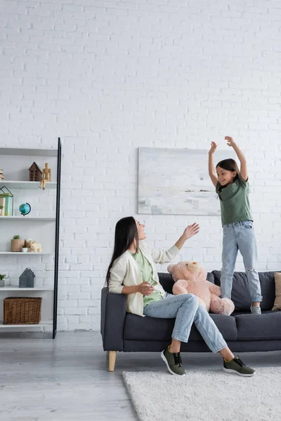 Niñera sorprendida mirando emocionada chica gesto mientras está de pie en el sofá en la sala de estar - foto de stock