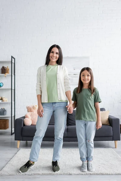 Full length of cheerful woman and girl standing and holding hands in modern living room - foto de stock