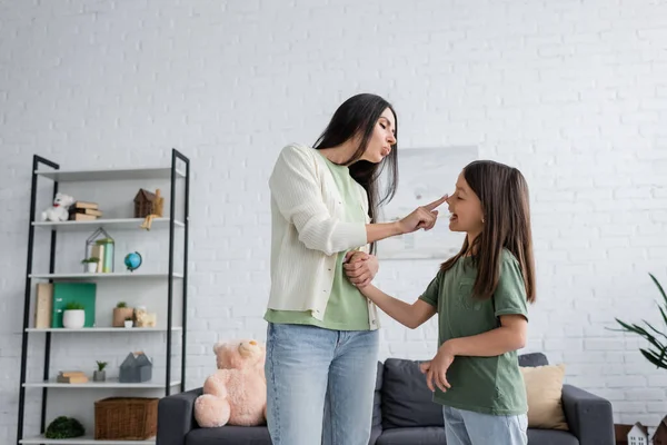 Funny babysitter pouting lips while touching nose of happy girl in living room — Stock Photo
