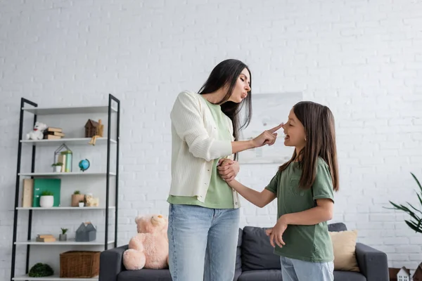 Funny babysitter pouting lips while touching nose of happy girl in living room — Foto stock