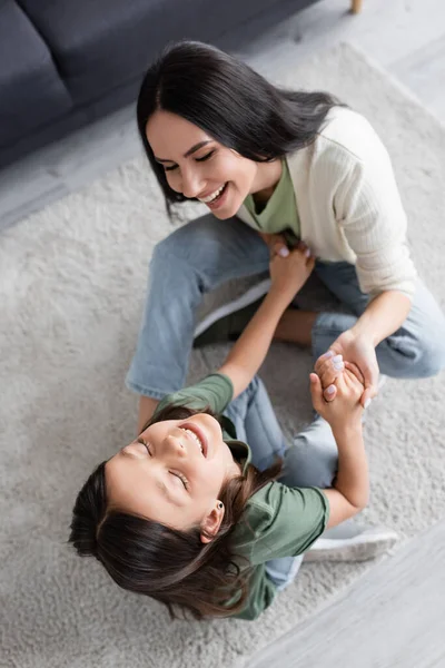 Top view of cheerful babysitter and girl having fun on carpet at home - foto de stock