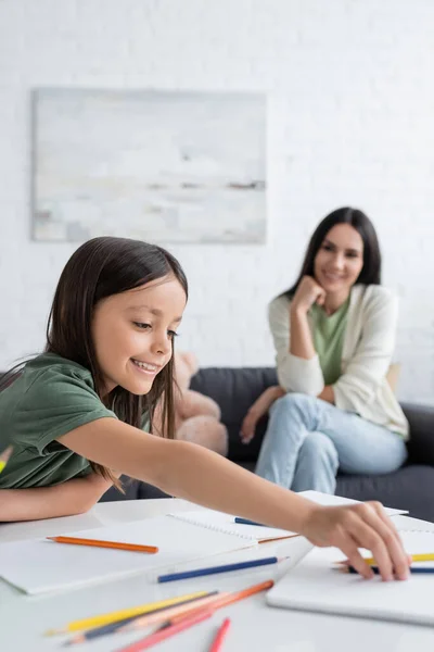 Fille heureuse atteignant crayons colorés près de papiers sur la table et baby-sitter floue sur le fond — Photo de stock