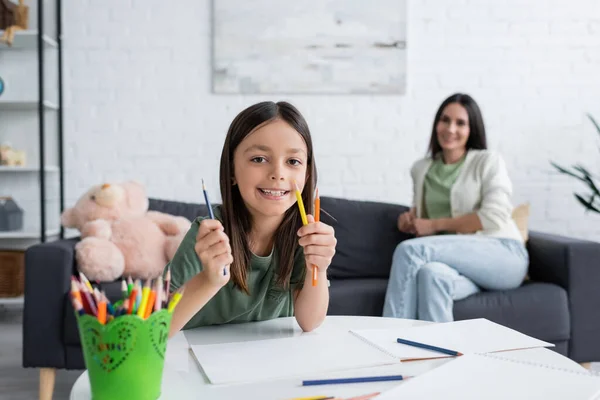 Menina alegre segurando lápis coloridos perto de papéis na mesa e babá borrada no fundo — Fotografia de Stock